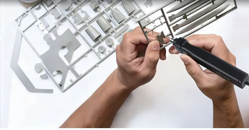 Hands trimming a small model part beside a plastic sprue frame